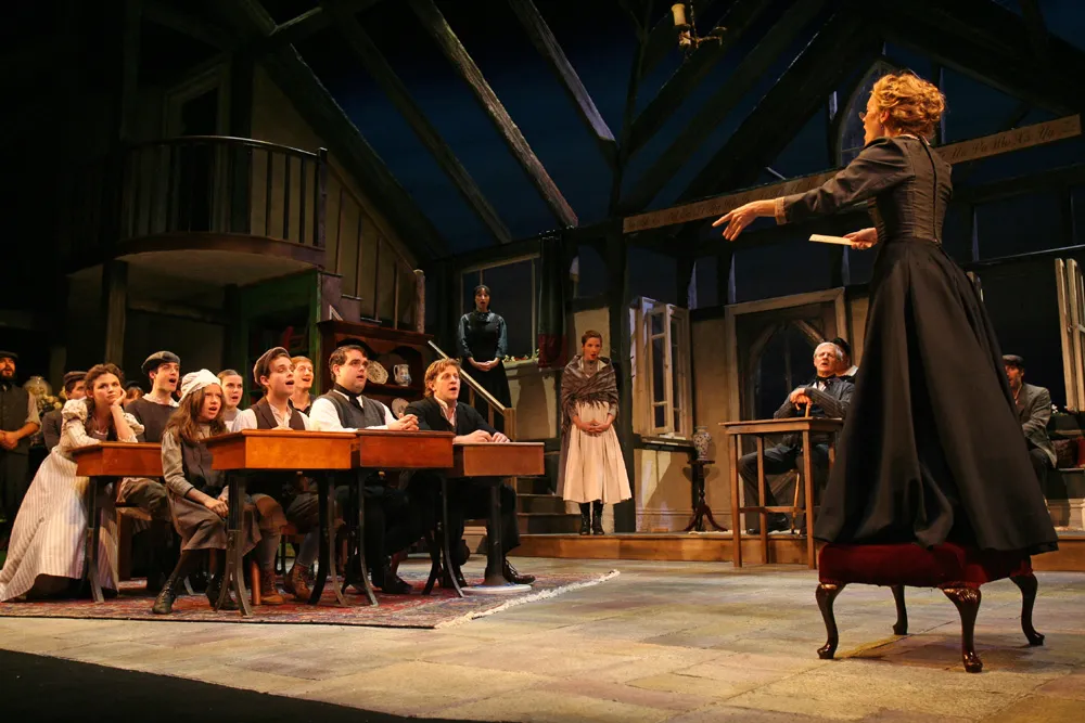 During a play, an actor sits at the side of a table with a group of people looking on. A woman stands up to address them. The room is dark with wooden floors and walls.