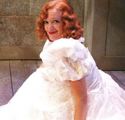 A woman sits on the ground wearing a white dress with lace trim. The dress reaches her ankles. Her hair is curly and red. She has blue eyes and a smile.
The wall behind her is stone. There is a closed door on it. A window is next to the door.