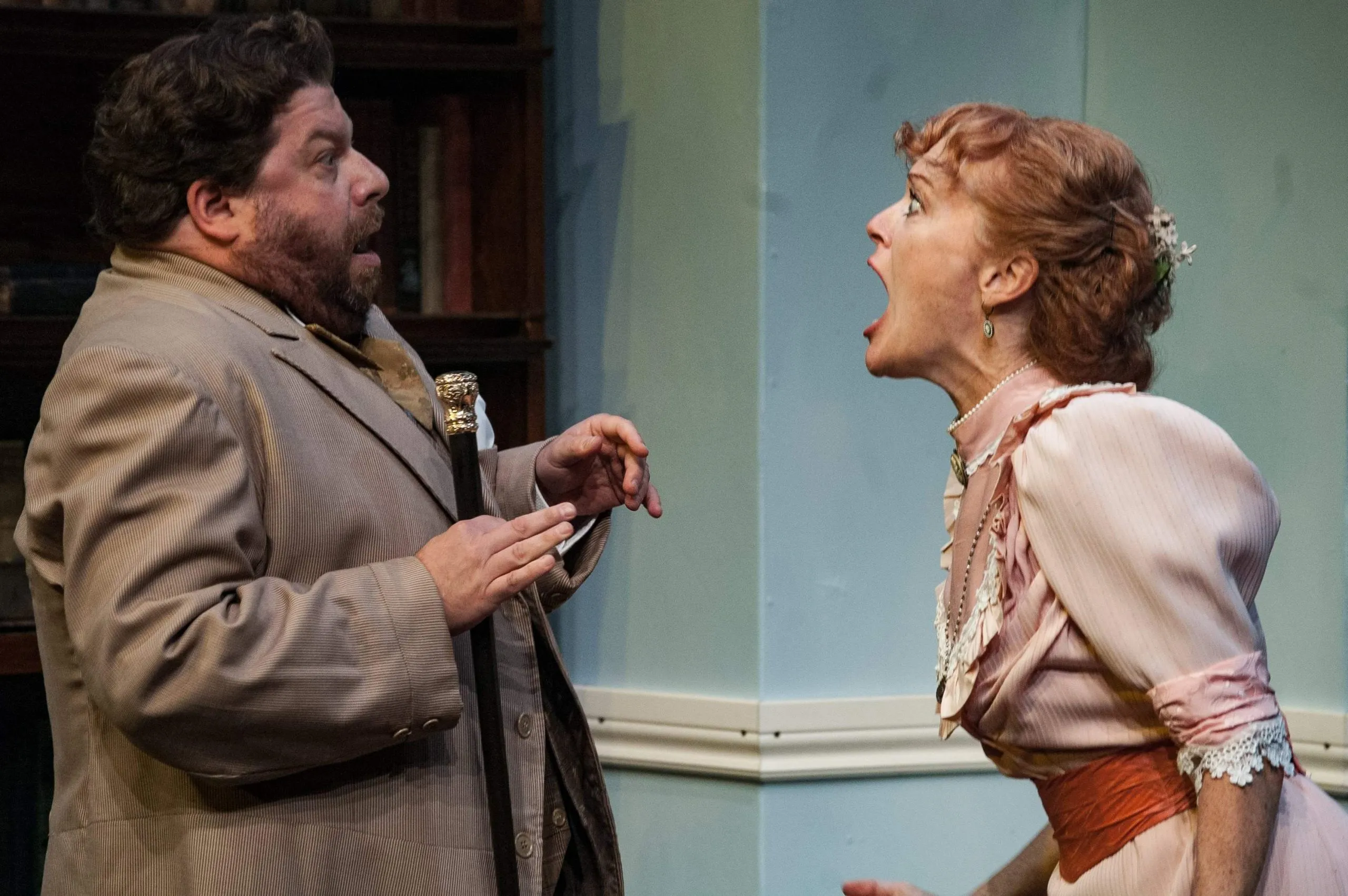 A man with a beard stands next to a woman wearing a pink dress with red sleeves while the woman opens her mouth as if she is going to scream or sing at the top of her lungs.