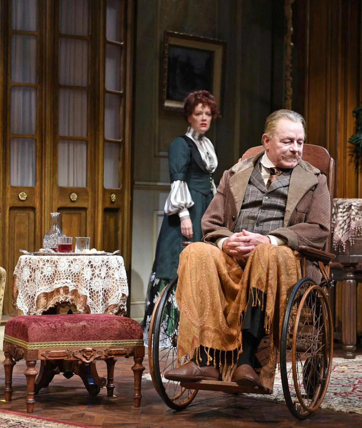 An elderly man with a cane is sitting on a wooden chair in the room with his wife who has her hand on his leg. The two of them are smiling at each other while they sit by the table that is covered with a white tablecloth and has candles on it. The man is wearing a brown sweater vest over a plaid shirt and is sitting in a wheelchair. In the background, there's a window with shutters behind them along with a painting of a boat hanging on the wall.