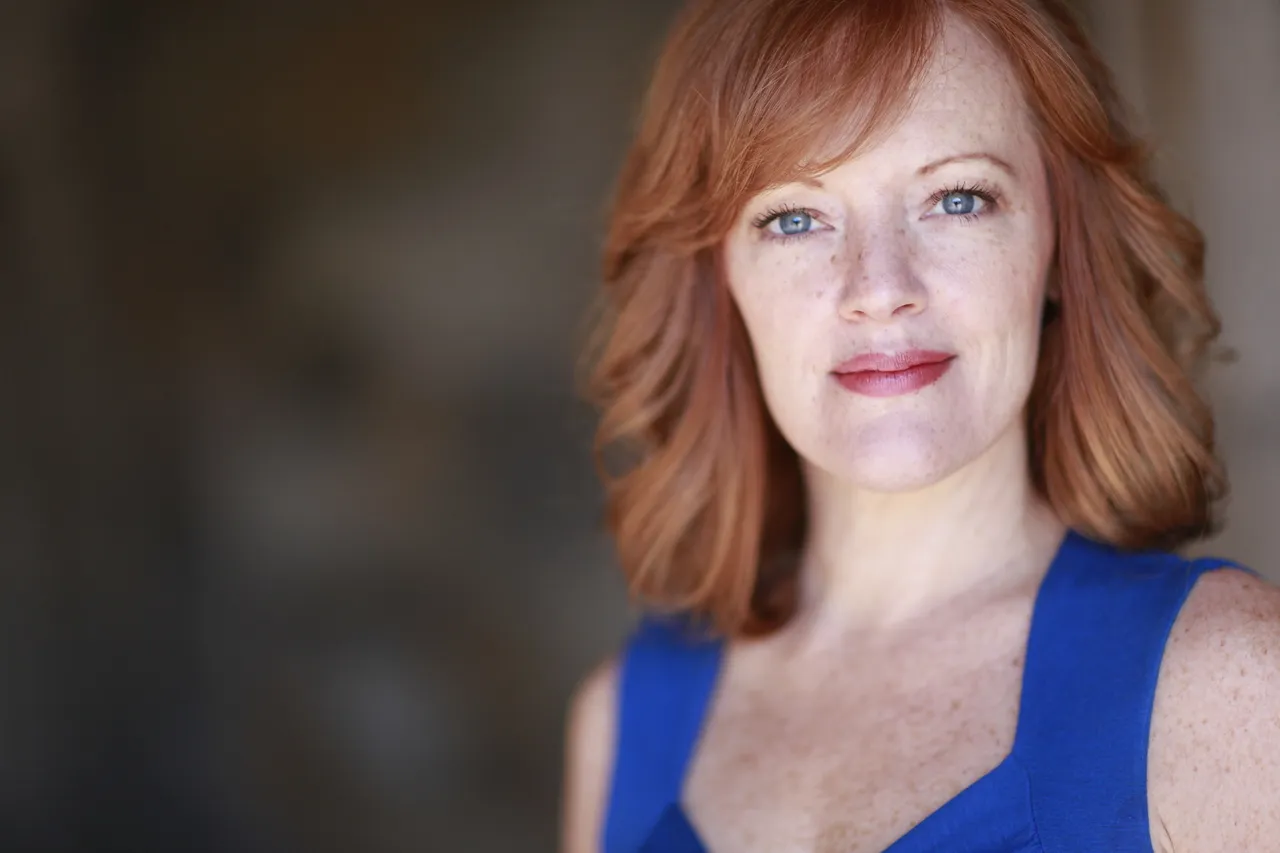 A woman with red hair poses for the camera. She is wearing a blue tank top.
