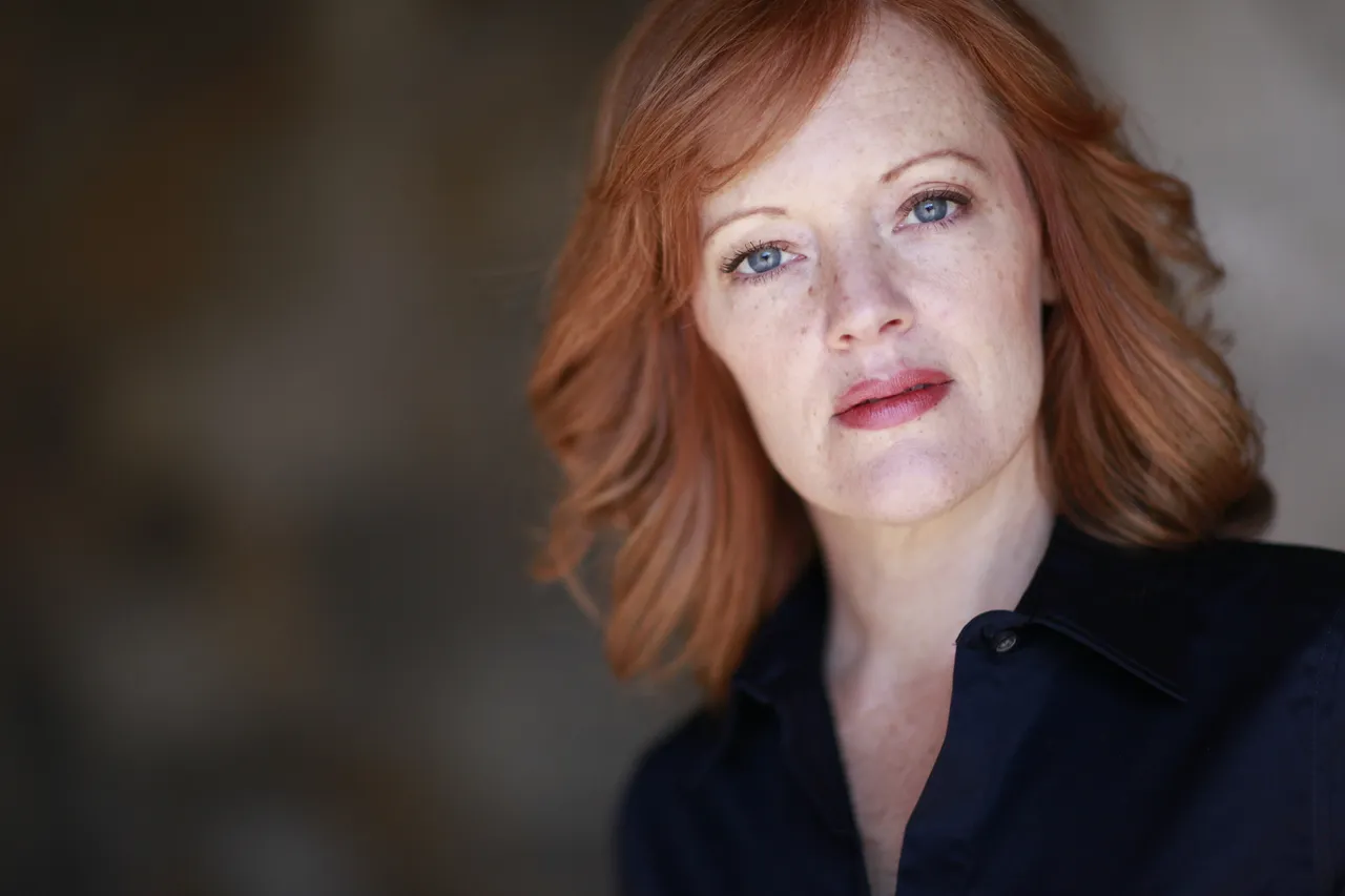 1. A woman with red hair is wearing a black shirt.
2. The blurred background behind the woman has an indistinguishable shape to it.