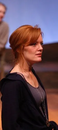 A woman with blonde hair wearing a dark blue sweater stands on a stage under the spotlights.