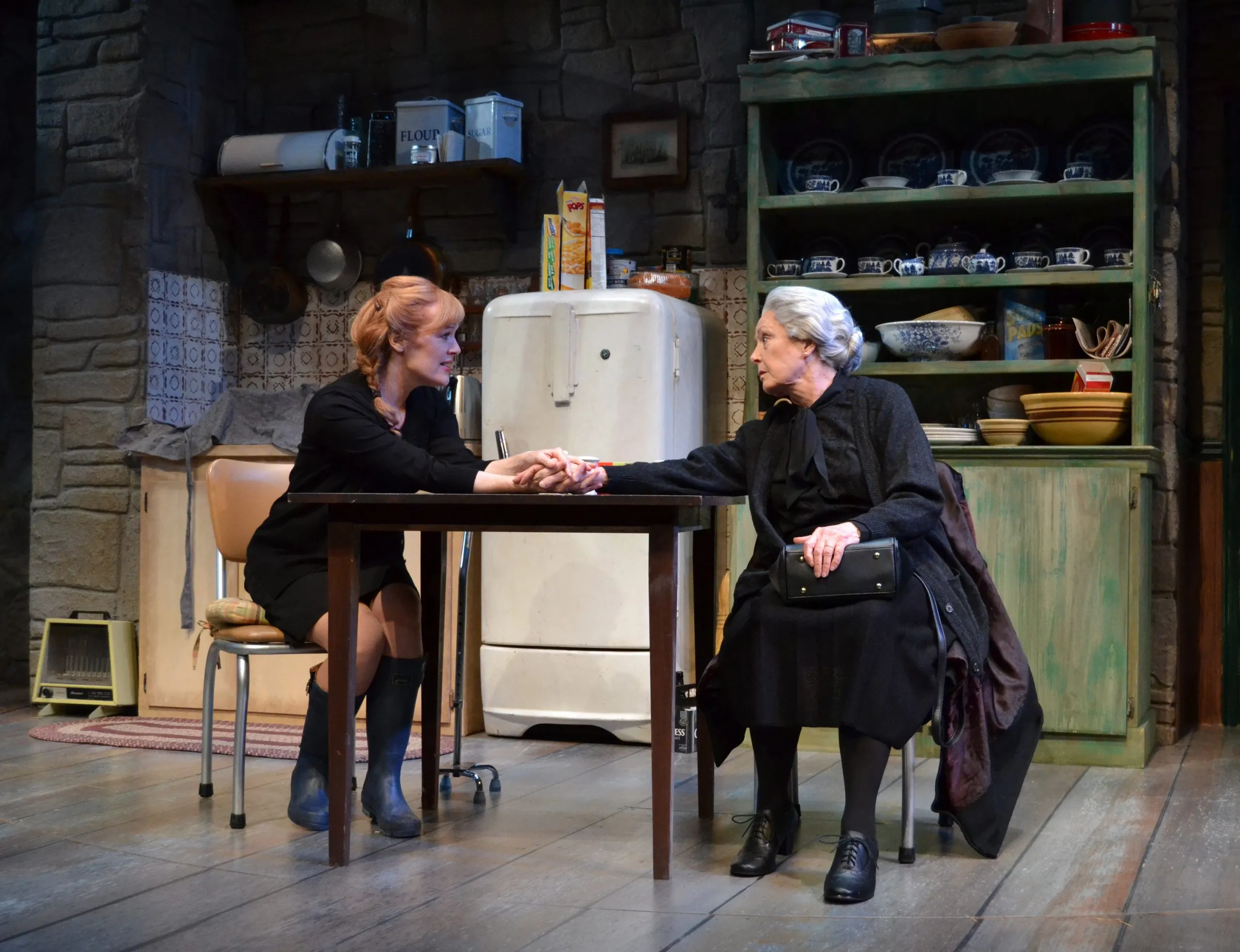 1) a woman with short red hair sitting at a table shaking hands with another woman with long white hair.
2) Woman with short blond hair and black dress.
3) A woman wearing a dark sweater is smiling.
4) A refrigerator, dishwasher, oven, and sink are behind the women.
5) Shelves filled with bowls, plates and other objects can be seen in the background.