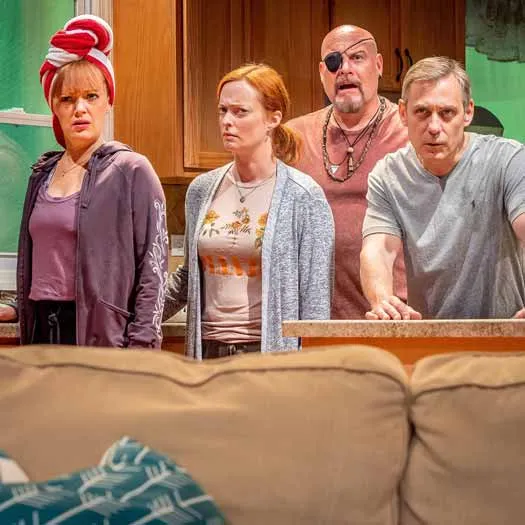 A scene from a TV show with four people standing behind a couch. The woman on the far left is wearing a red and white hat. The man in the center has his eyes closed and appears to be deep in thought. One of the men is balding, while another has a goatee. A wall clock can be seen in the background, with its hands indicating that it's nearly 12 o'clock.