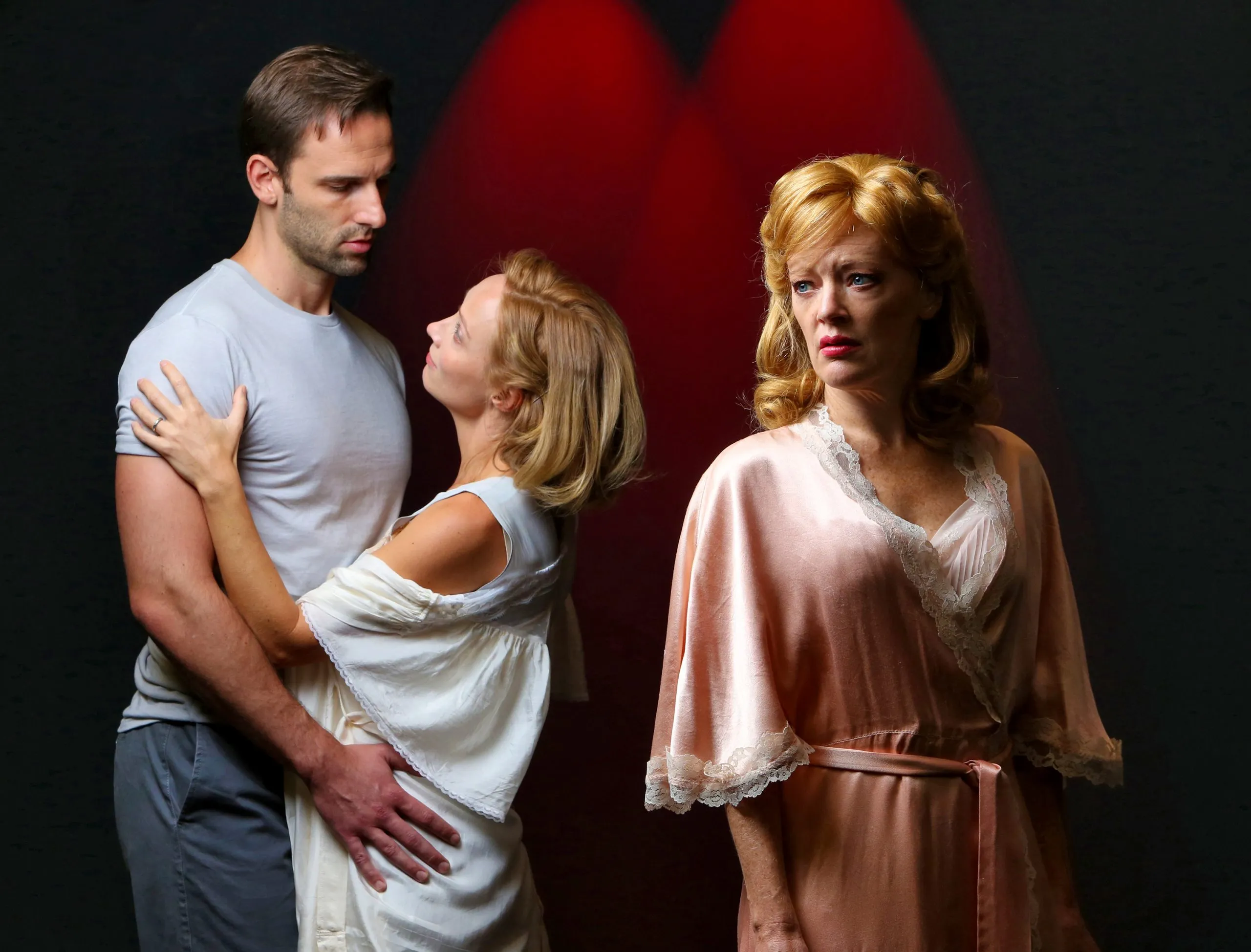 * A man wearing a gray t-shirt is hugging a woman with blonde hair who has her hand on his shoulder.
* The third person, a woman with red hair, stands behind them both in a pink dress.