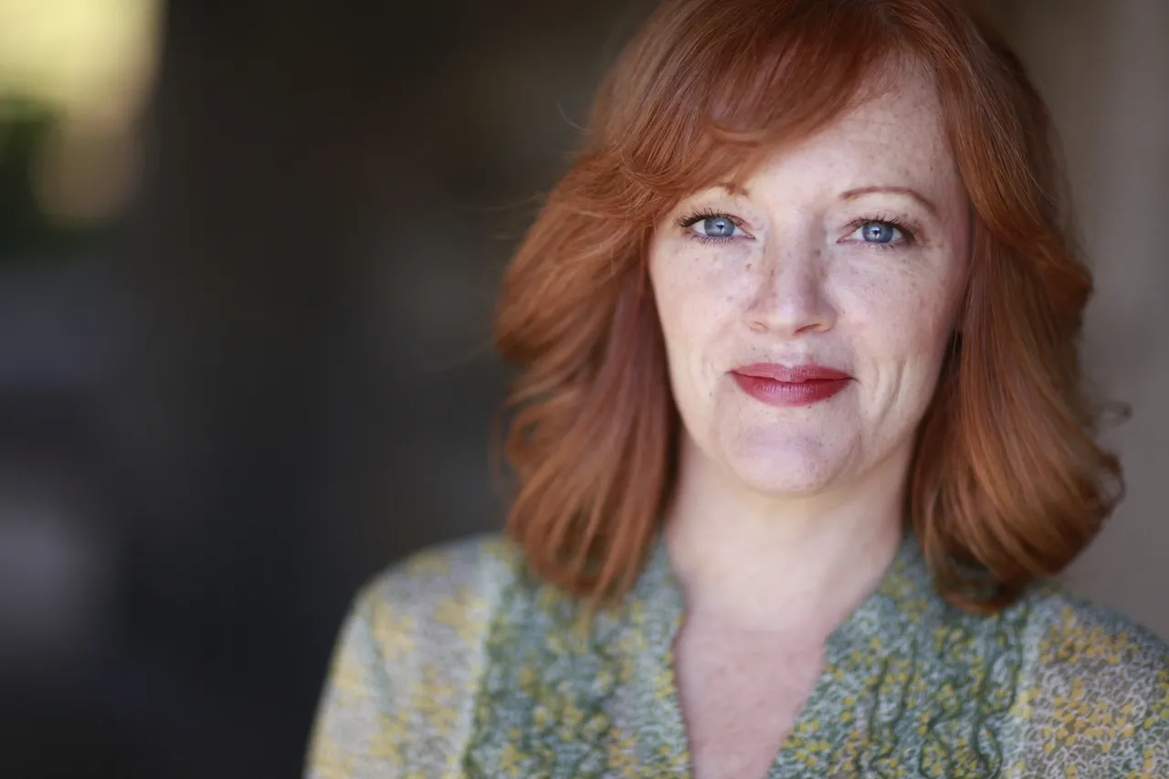 The image features a woman with red hair that is colored red on the ends, wearing a blue shirt. She appears to be smiling at the camera. The background of the photo is out of focus but it looks like there might be an alleyway or walkway behind her.