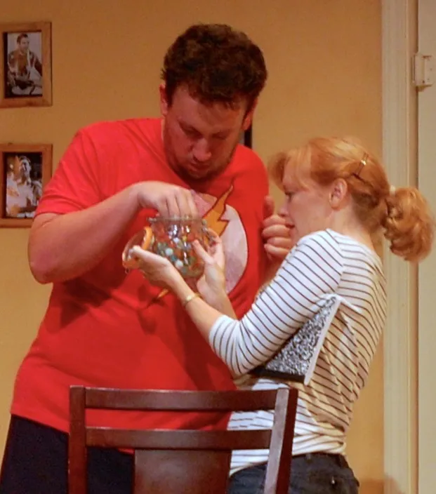 A man with a red shirt is holding a glass bowl filled with small gems. A woman wearing a striped shirt is looking at the man, seemingly interested in the jewels inside the bowl. The man appears to be presenting or offering something to the woman. There are framed photos hanging on a wall behind them and a chair can be seen as well.