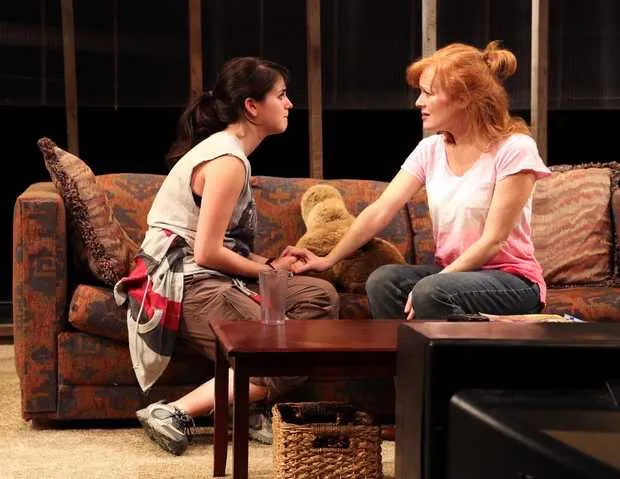 A woman wearing a red pink shirt is sitting on the couch with another woman who is holding her hand. There's a brown teddy bear next to them. A small table beside the couch has a glass of water, and there's a wicker basket underneath it.