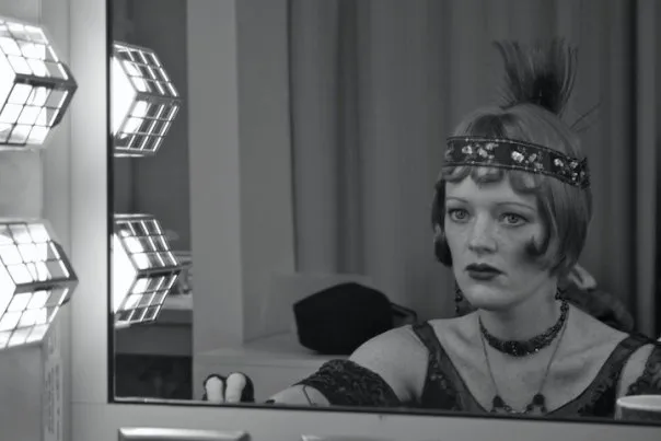 The woman is wearing a feathered headdress. She looks somber as she gazes at her reflection in the mirror. The photo is black and white, giving it an antique feel. A man's tie can be seen on the bottom right of the image.
