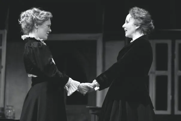 Two women are shaking hands while one woman behind them watches. One of the women has grey hair. This is a black and white photo. The two women are wearing dresses with ruffled sleeves. The left hand of each woman holds another women's hand as they shake.