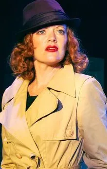 A woman wears a tan trench coat and hat, standing under blue lights.