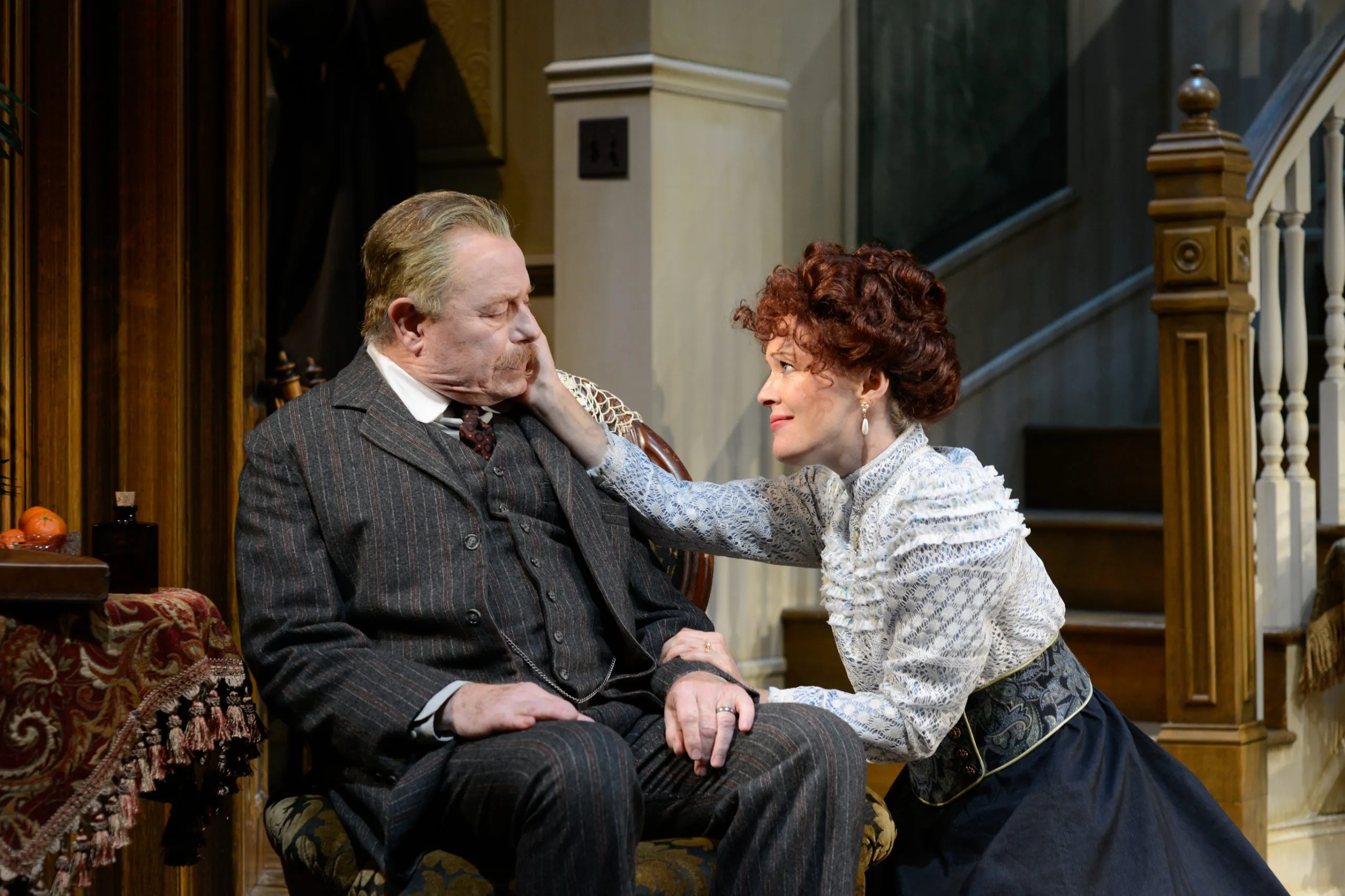 * A man with brown hair wearing a suit is sitting on a chair.
* A woman with red hair is touching his face.
* The scene takes place in a room with beige walls, white door frame and wooden staircase.